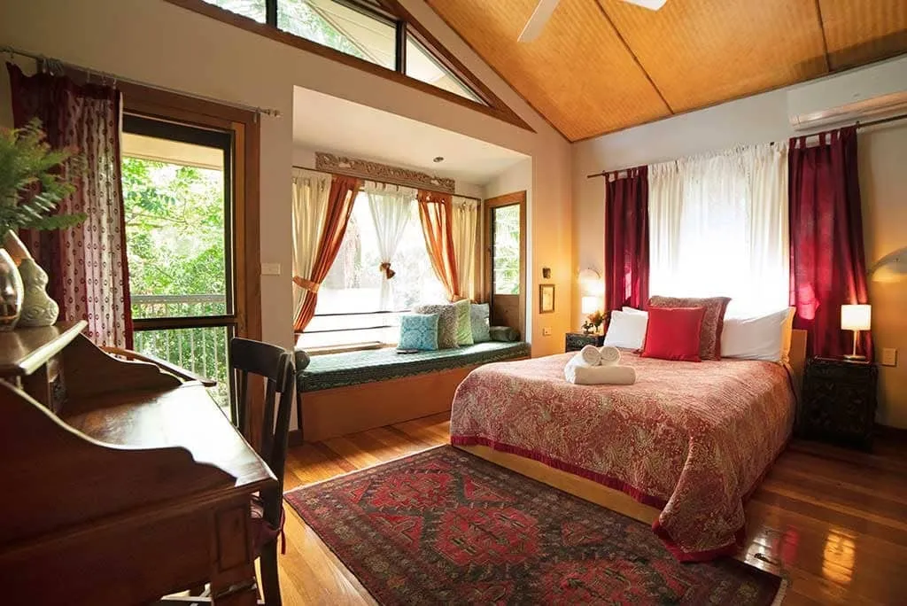 A guest bedroom at Sangsurya with a red quilted bed, carved desk and a window seat looking out to the trees